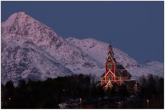 Bilde av Buksnes kirke i vinterlys med snøkledte fjell i bakgrunnen