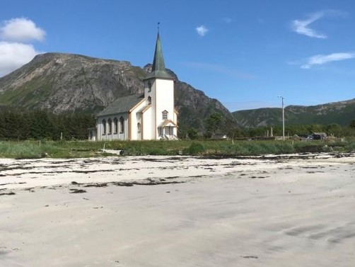 Sommerbilde av Valberg kirke, med blå himmel, stranda foran og fjell i bakgrunnen.